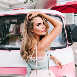 Chica rubia con mechas babylight, tocándose el pelo y posando delante de una furgoneta rosa vintage