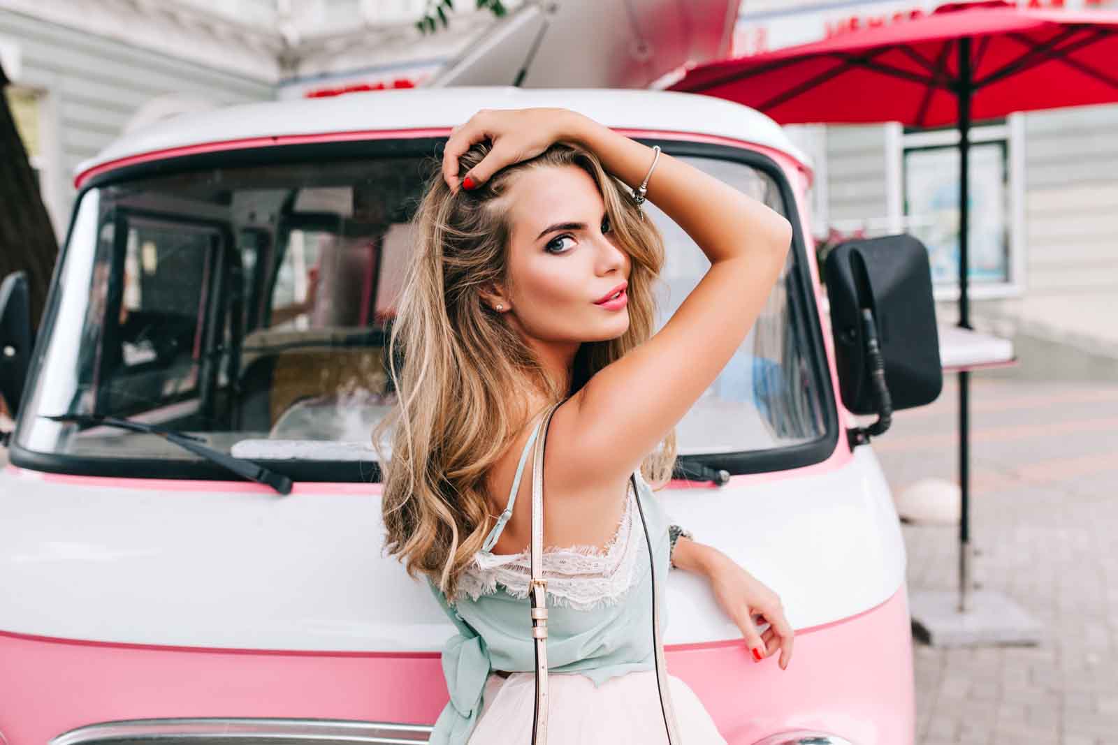 Chica rubia con mechas babylight, tocándose el pelo y posando delante de una furgoneta rosa vintage